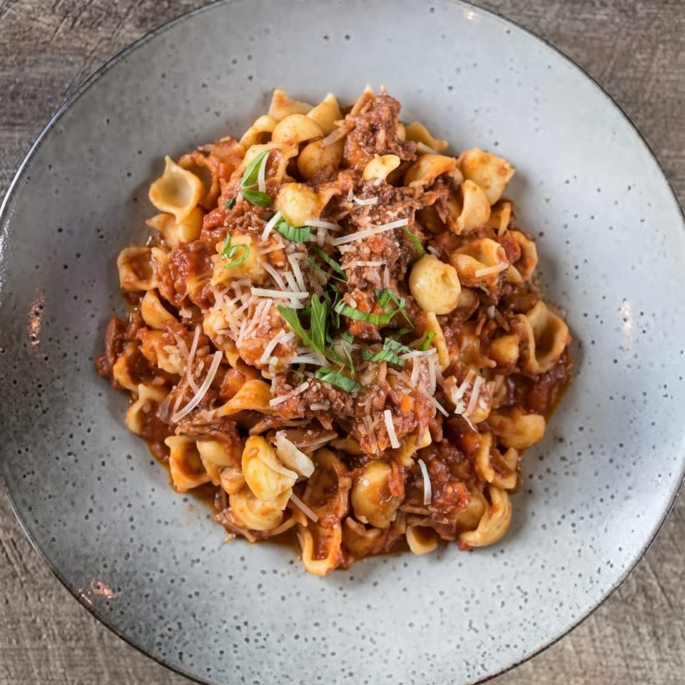 A close-up of orecchiette pasta with a rich red meat sauce, topped with grated cheese and fresh herbs.