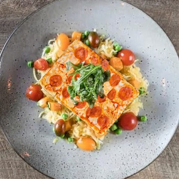 A square pepperoni pizza slice with arugula on a bed of pasta with tomatoes and peas.