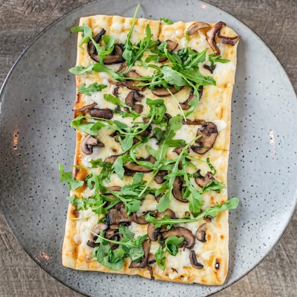 Rectangular flatbread pizza with melted cheese, sautéed mushrooms, and fresh arugula on a gray plate.