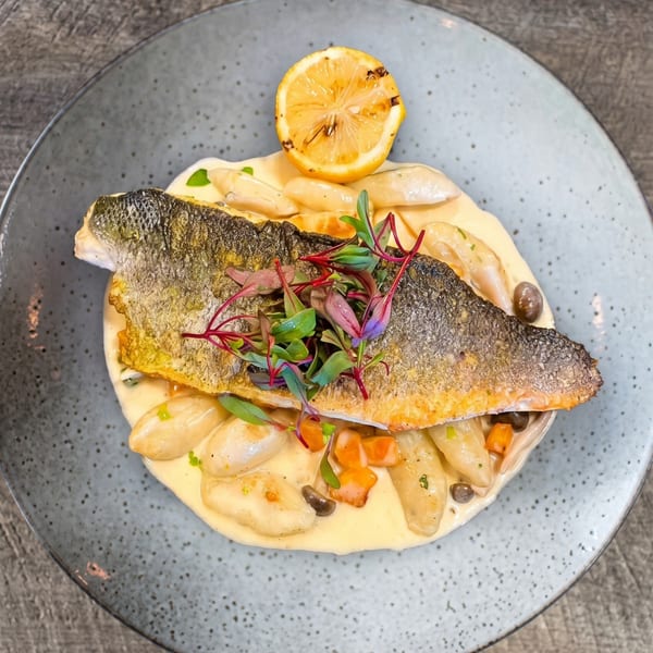 Pan-seared fish with crispy skin, gnocchi, capers, and microgreens, garnished with a grilled lemon.